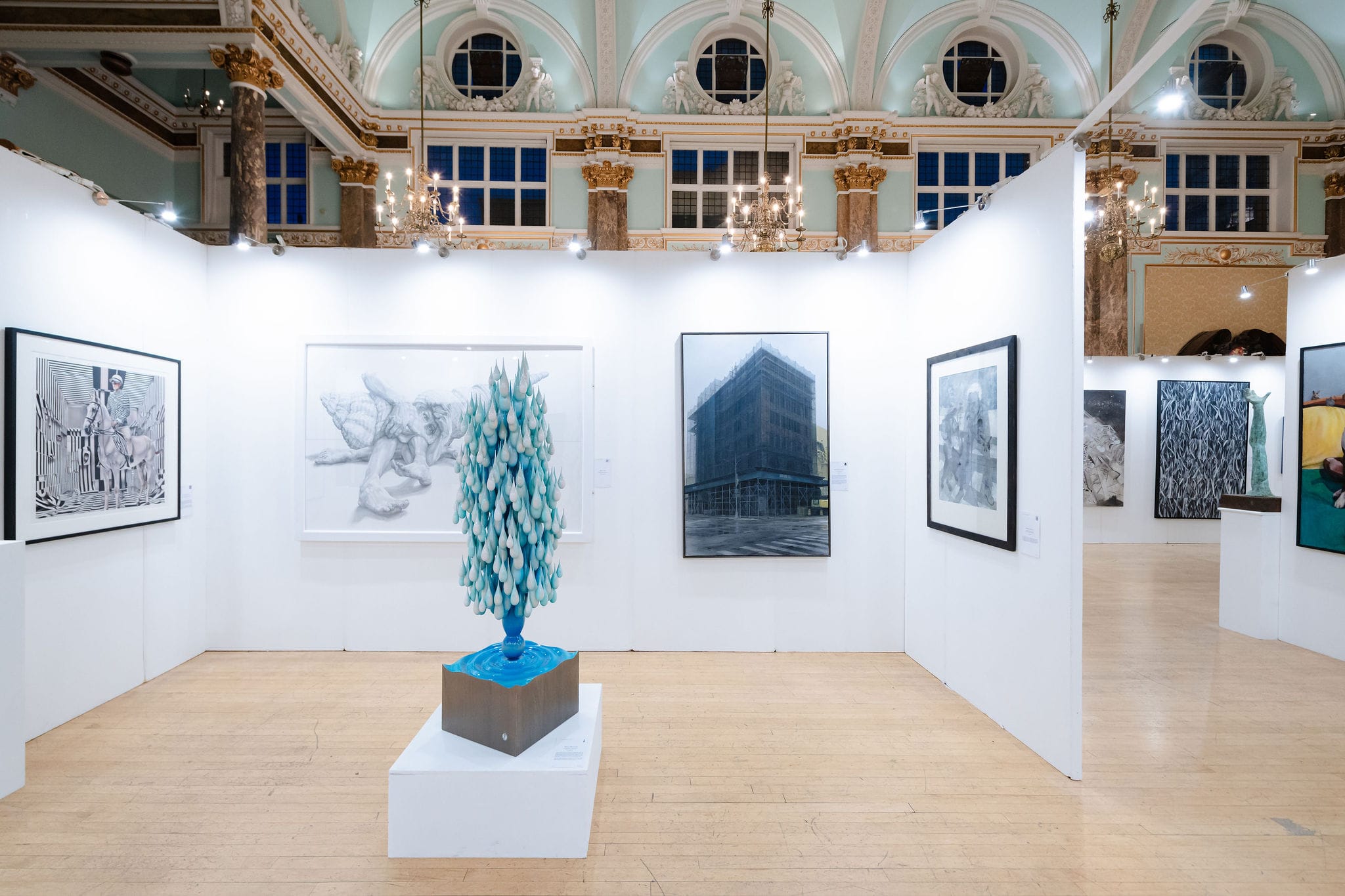 A wide view of the hang, a sculpture on its plinth at the centre beneath the hall's columns and chandeliers.