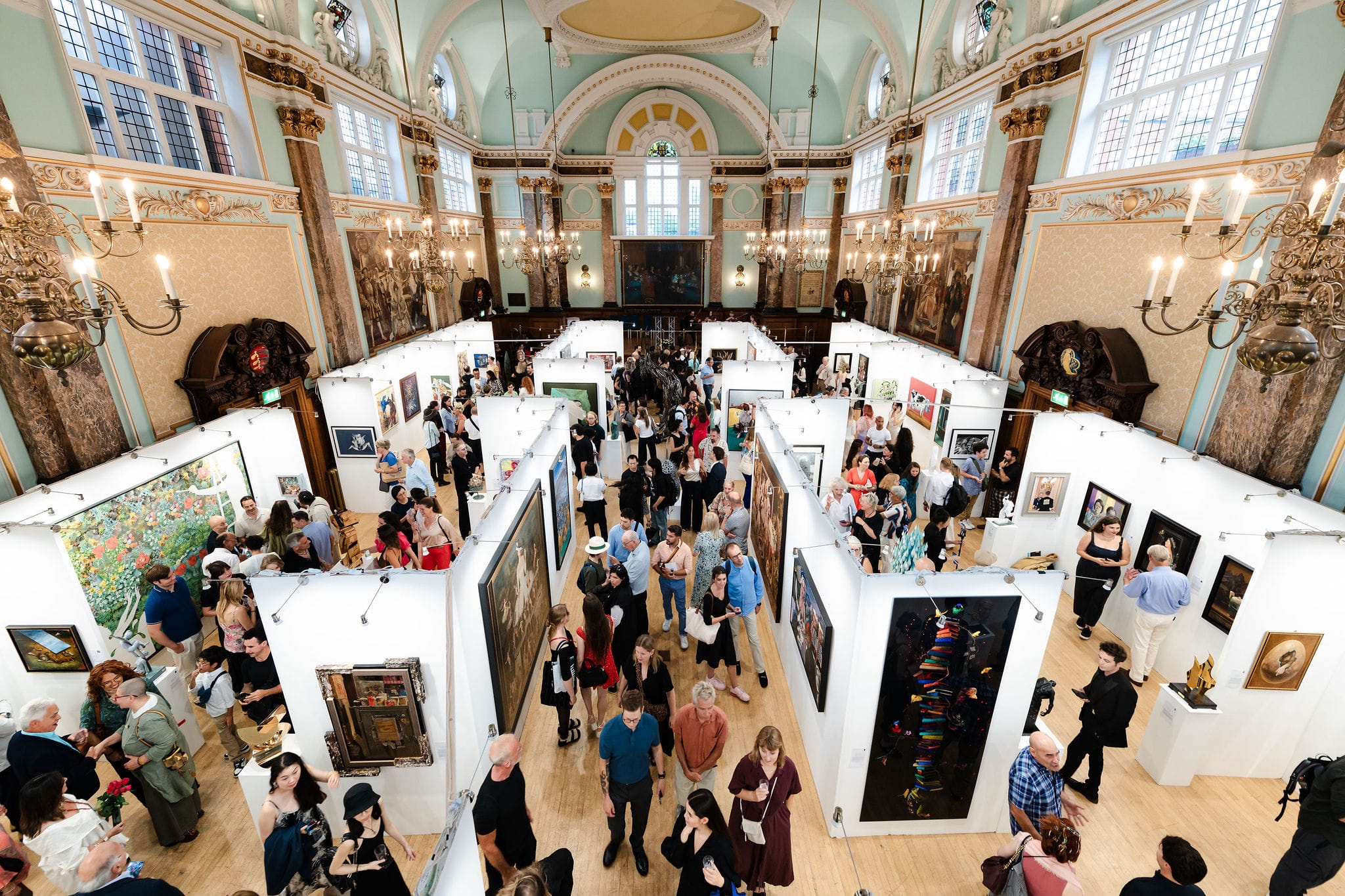 The exhibition seen from the balcony at the private view: temporary white walls hung with work laid out in a grid across the floor of the gilded hall, visitors moving between them.
