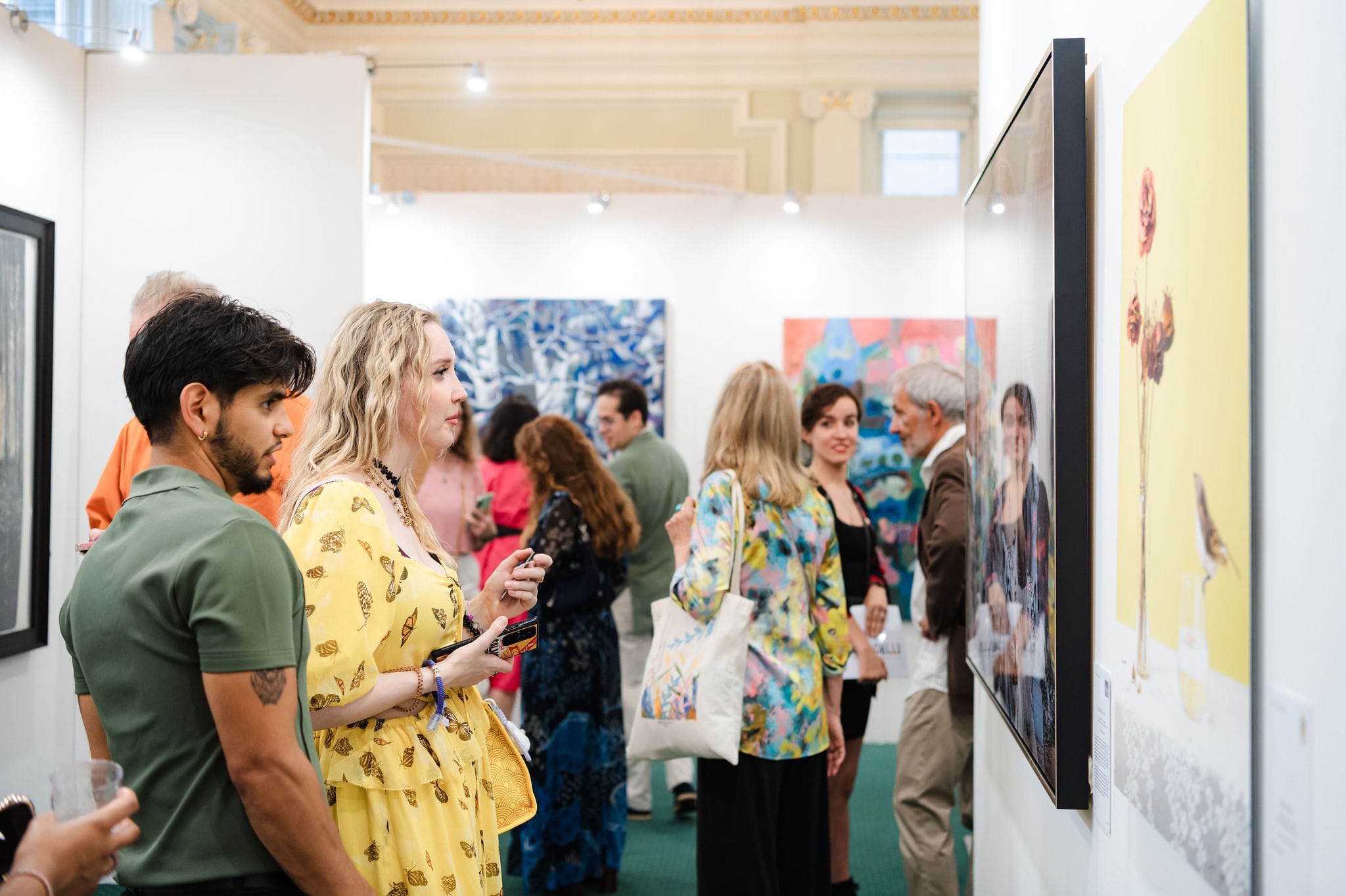 Guests talking among the paintings at the private view, a woman in a yellow dress at the front of the crowd.