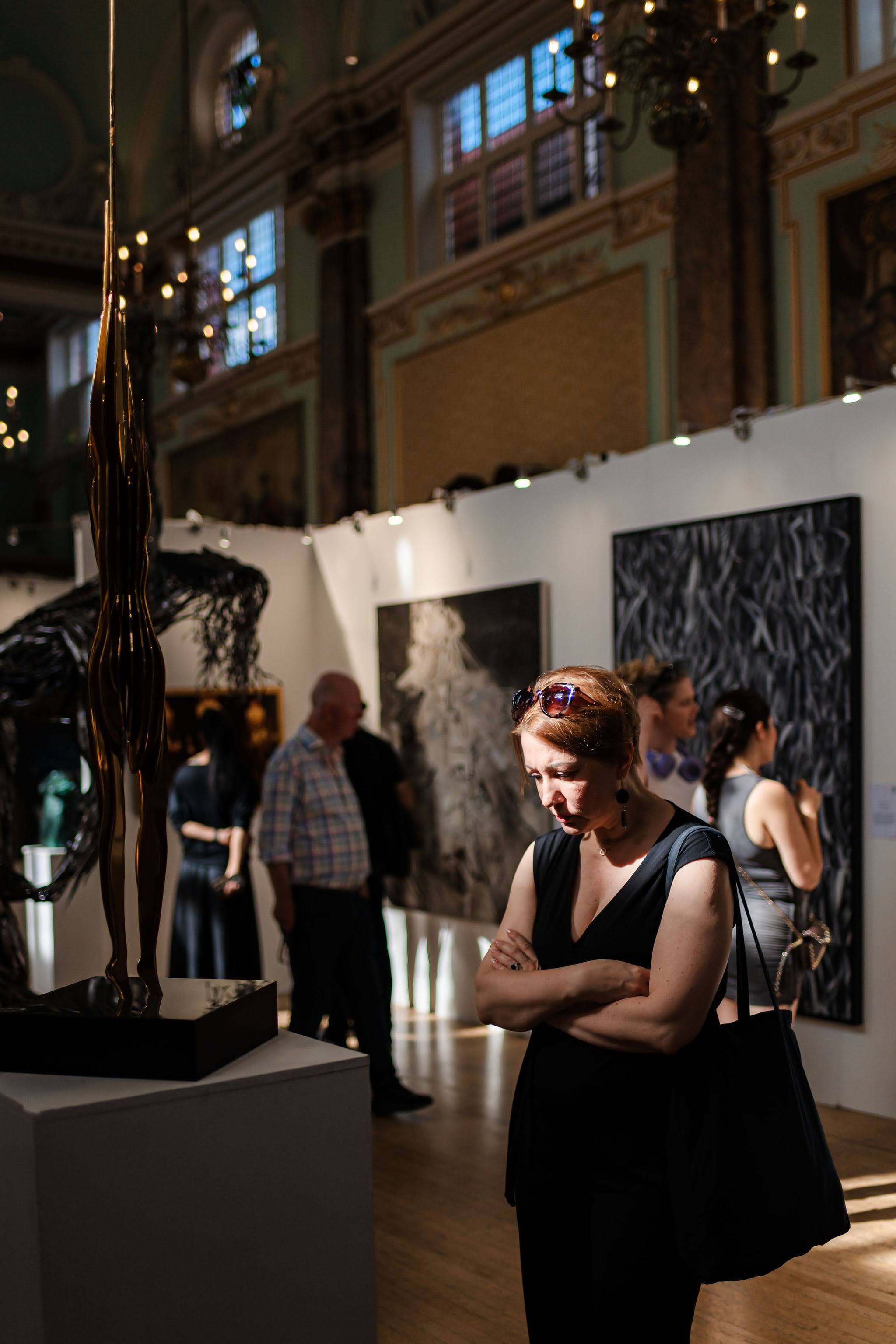 A visitor pausing beside a tall bronze figure, the hang and the lit hall behind her.